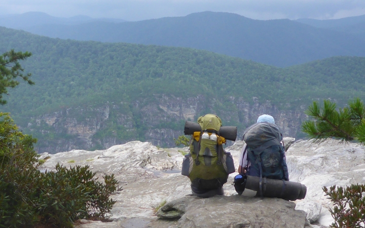Two people wearing backpacks sit on a rock, facing away from the camera. They are looking out over a vast and green mountainous landscape.